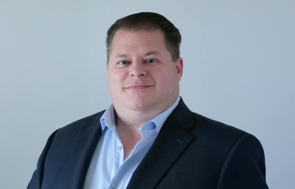 Chris Legg, a man with short, dark hair wearing a navy suit jacket and light blue shirt, stands in front of a plain light gray background, smiling slightly at the camera.
