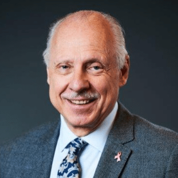 Smiling older man with grey hair and mustache wearing a grey suit, patterned tie, and a pink ribbon pin on his lapel, posed against a dark background. This is Stefan Glück.