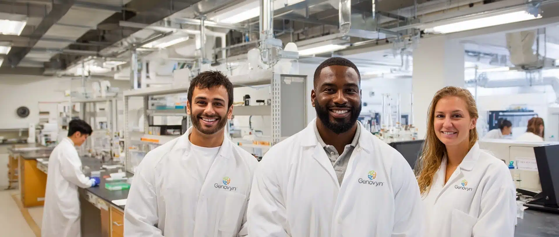 Three scientists in white lab coats stand smiling in a modern laboratory, with lab equipment and another person working in the background.