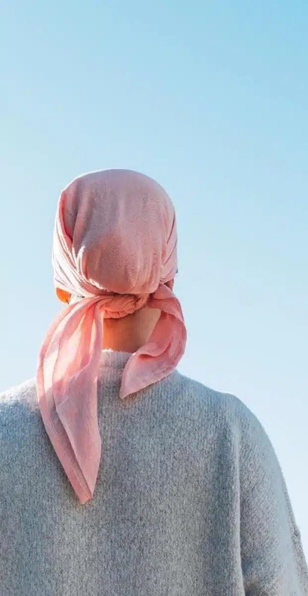 A person wearing a light pink headscarf and a gray sweater, seen from behind, stands against a clear blue sky.