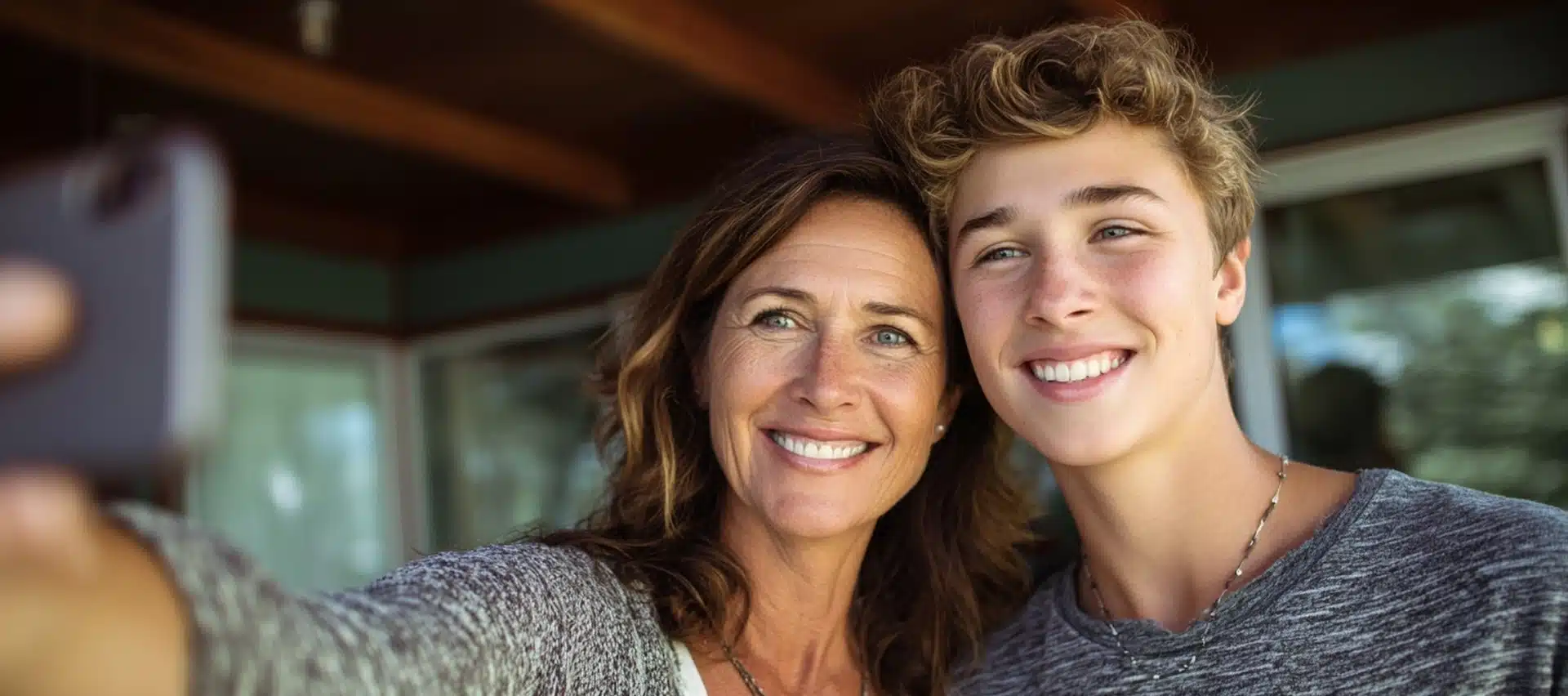A woman and a teenage boy smile while taking a selfie together indoors, with natural light coming through windows in the background.