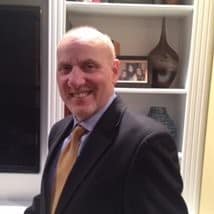 Dr. David Levine, a smiling man in a suit and tie, stands indoors in front of white shelves adorned with decorative vases and a framed photo.