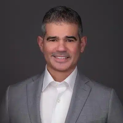 Claudio Faria, a man with short gray hair wearing a light gray suit jacket and white shirt, smiles at the camera against a dark gray background.