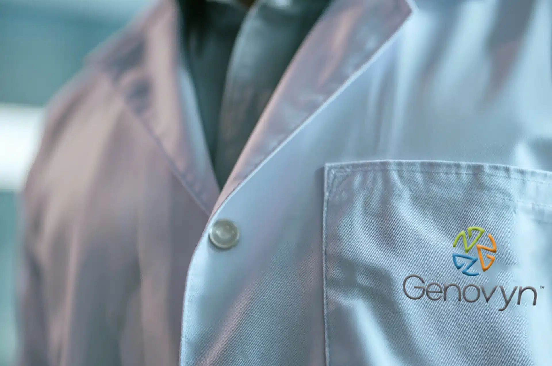 Close-up of a person wearing a white lab coat with the Genovyn logo and colorful emblem embroidered on the chest pocket.