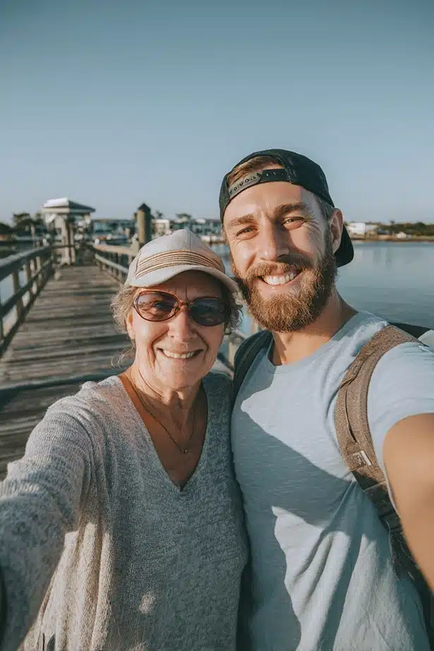 A smiling older woman and a bearded younger man take a selfie together on a wooden dock by the water, with clear skies and buildings in the background.