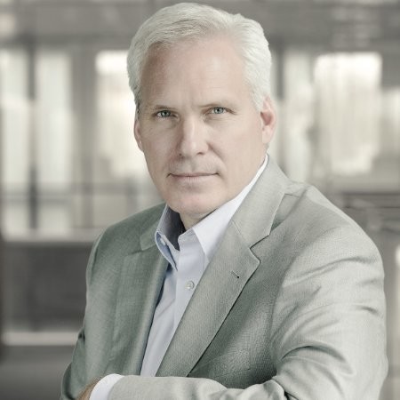 A mature man with short white hair, identified as Jeff Bagshaw, wears a light gray suit jacket and light blue shirt, posing while seated in a modern, blurred office setting. He looks directly at the camera with a neutral expression.