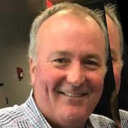 A middle-aged man with short light hair, identified as Jeff Reiniche, smiles at the camera. He is wearing a checked shirt indoors, with part of another person's face partially visible on the right side.