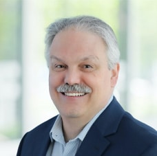 A middle-aged man with gray hair and a mustache, wearing a dark blazer and light blue shirt, smiles at the camera against a blurred, bright background.