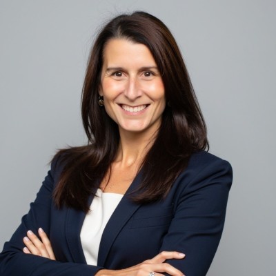 Sare Largay, a woman with long dark hair, wears a navy blazer over a white top and stands smiling with her arms crossed against a plain light gray background.